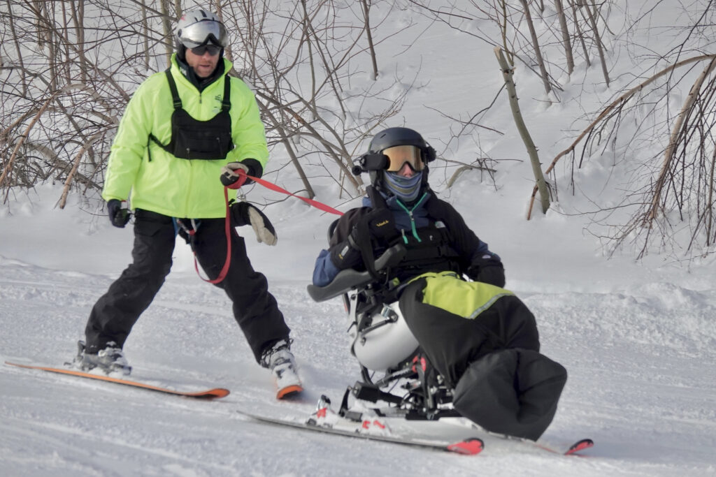 An athlete controls the TetraSki with the use of joystick, and is supported by an instructor with a tether.